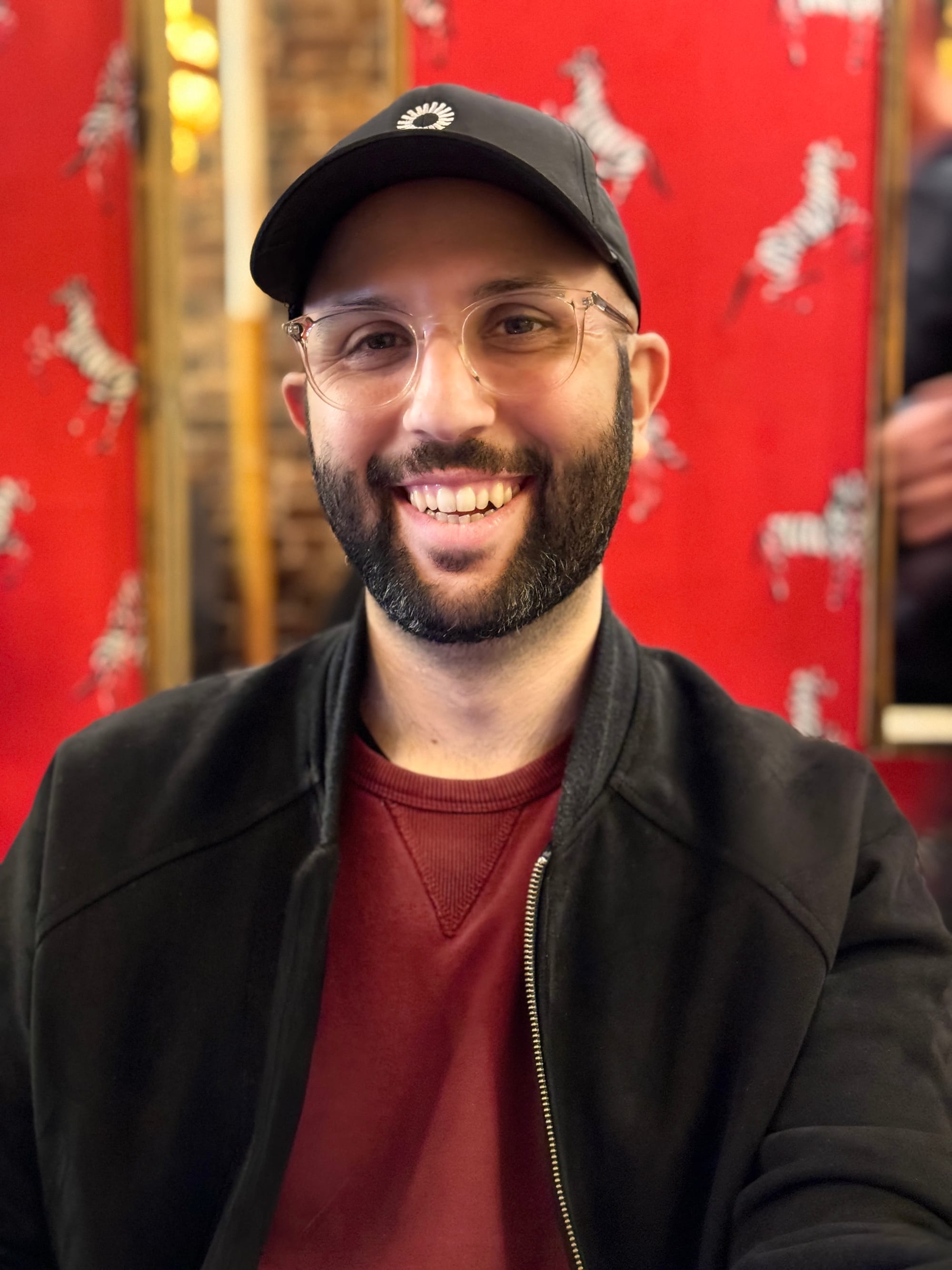 portrait photo of me, a smiling bearded white man in a red sweater, a black bomber jacket, and a black baseball cap
